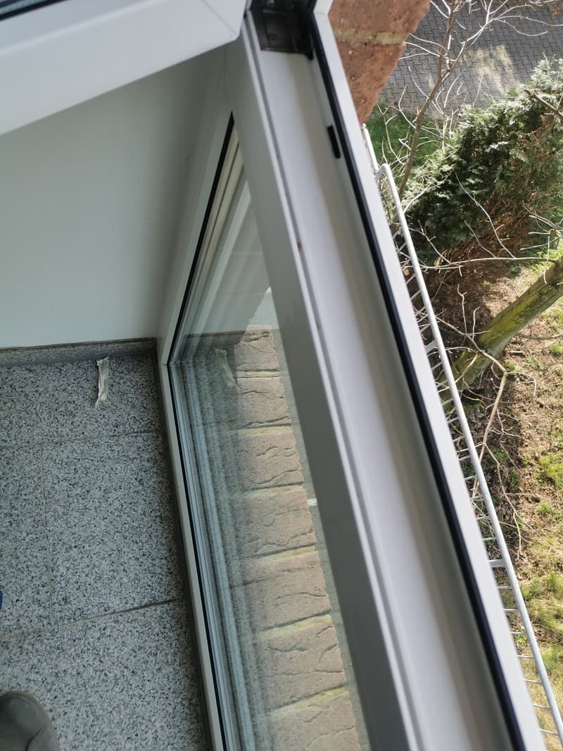 Ein Blick aus einem Fenster auf einen Balkon mit einem Baum und einer Mauer im Hintergrund. Das Fenster ist teilweise geöffnet.