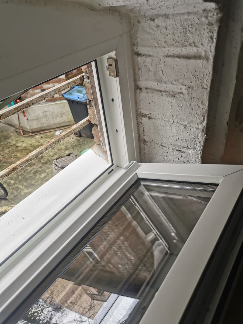 Ein offenes Fenster mit Blick nach draußen auf einen Garten mit einer Mülltonne und einem Korb. Das Fenster ist weiß und der Rahmen ist aus Kunststoff.