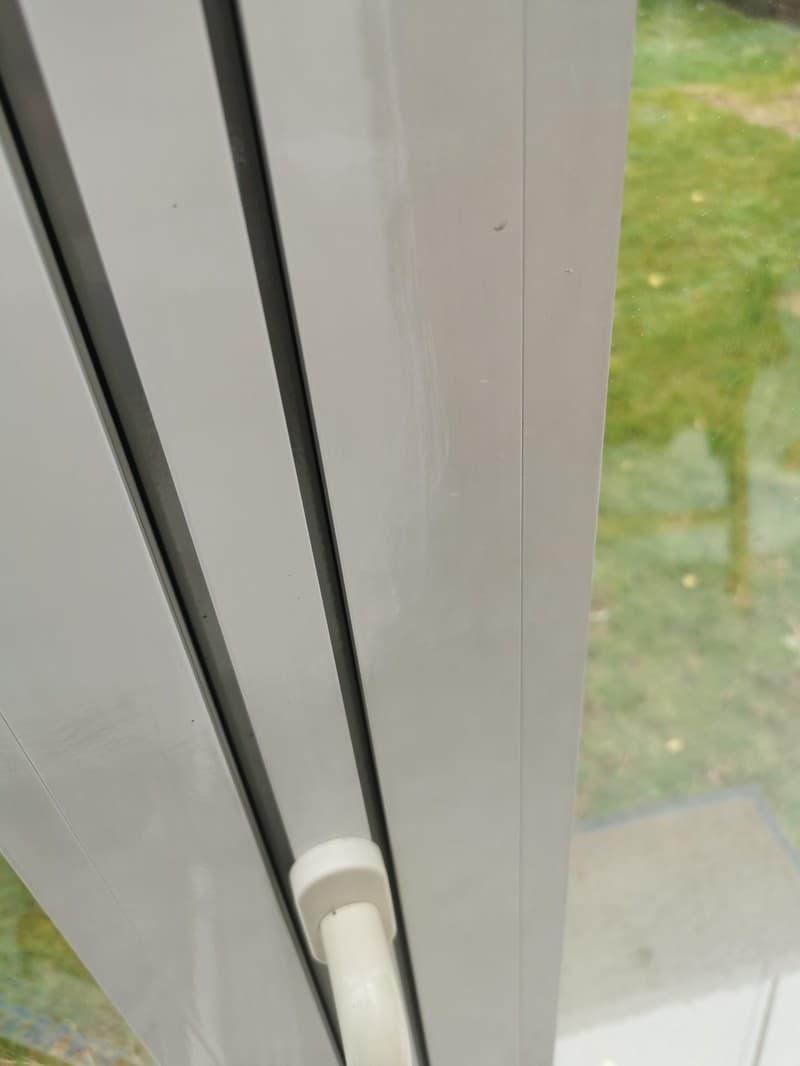 Nahaufnahme eines weißen Fensterrahmens mit Griff, der einen Blick ins Grüne ermöglicht.